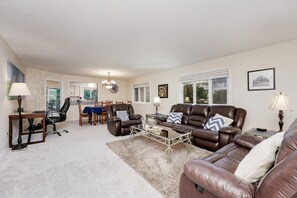 A living room with a brown leather sofa, a dining table with chairs, a chandelier, and a large window.