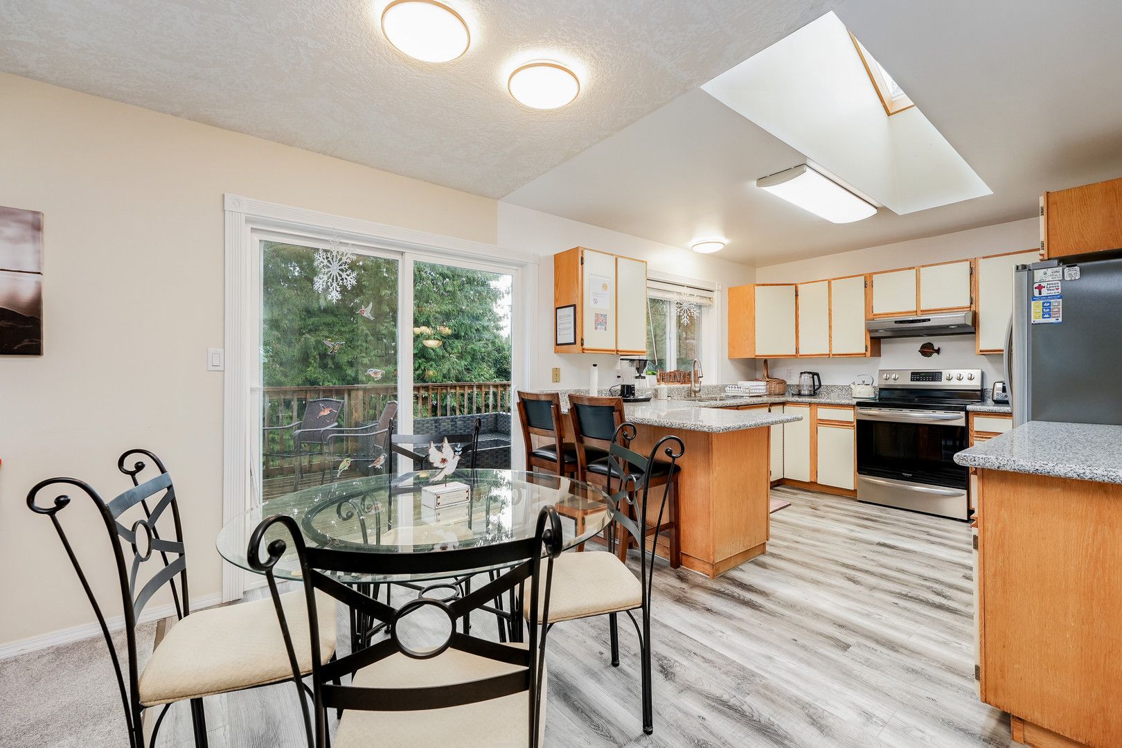 A modern kitchen with a dining area, featuring a glass table and chairs, wooden cabinets, and a large window overlooking a garden.