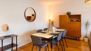 A dining area with a wooden table set for six, surrounded by chairs. A mirror hangs on the wall above the table, and a cabinet with a radio is to the right. - Le ViLoNa - T3 moderne avec parking privé (Rennes)
