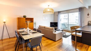 A modern living room with a dining area, a sofa, a TV, and a large window. - Le ViLoNa - T3 moderne avec parking privé (Rennes)