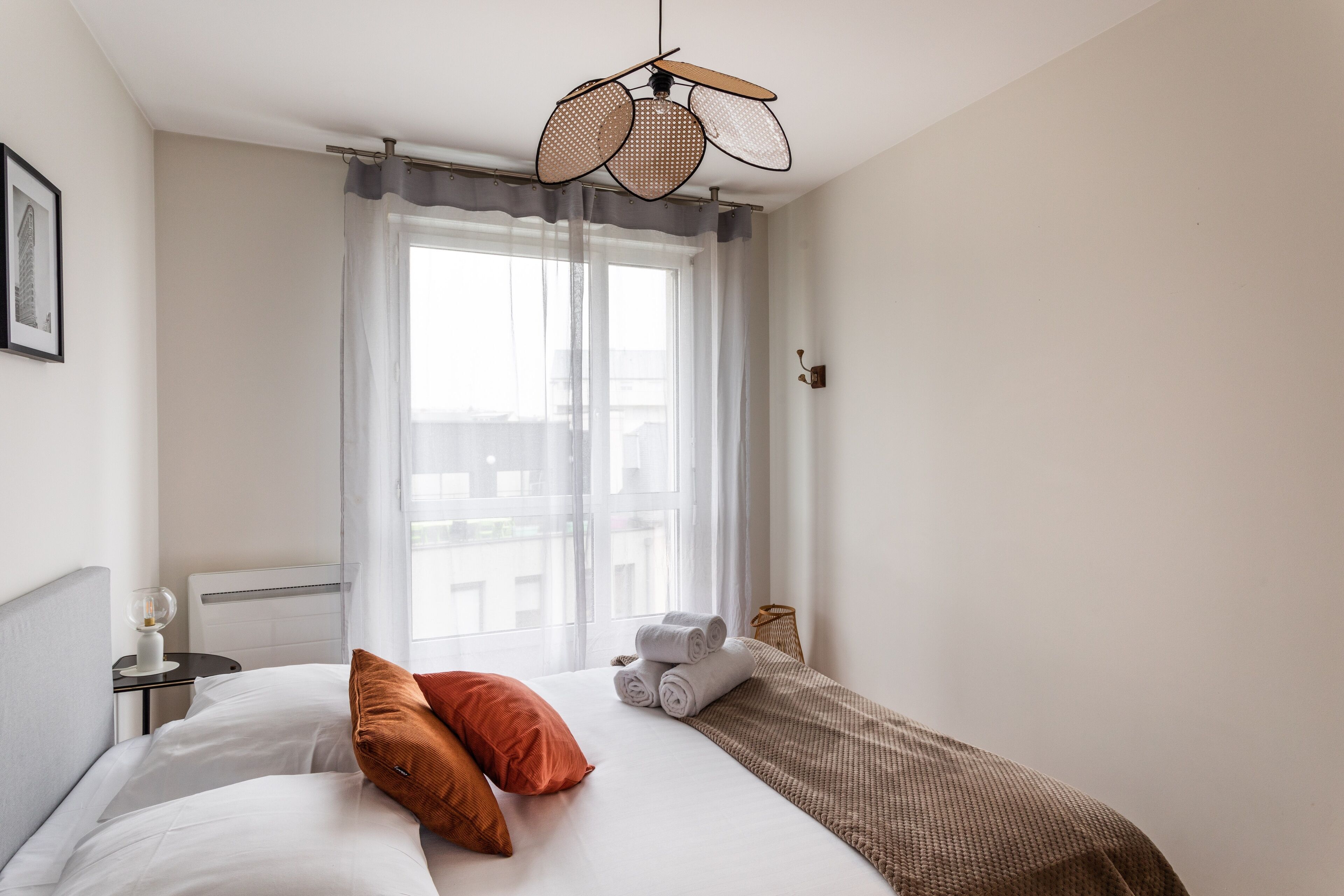 A hotel room with a bed, a bedside table with a lamp, a window with sheer curtains, and a view of buildings outside.