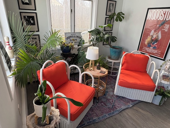 A cozy seating area with wicker chairs and a red cushion, surrounded by various potted plants and framed pictures on the walls. - Waterfront Oasis w/ Great Views, 5 beds & deck over the Creek! (Saugerties)