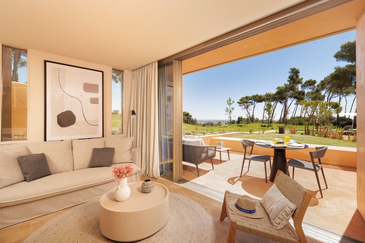A modern living room with a sofa, a coffee table, and a dining area with chairs and a table. There is a large window offering a view of a garden and trees.