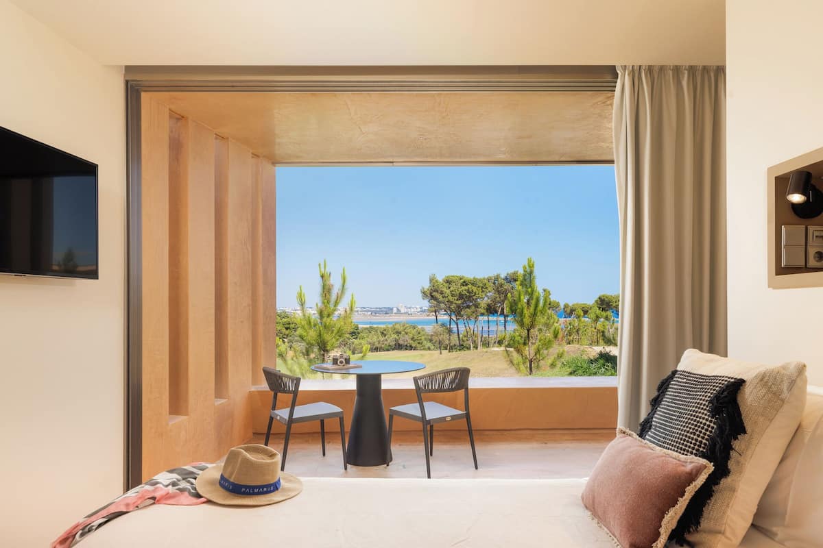 A hotel room with a balcony, a table, two chairs, a hat, and a bed with pillows.