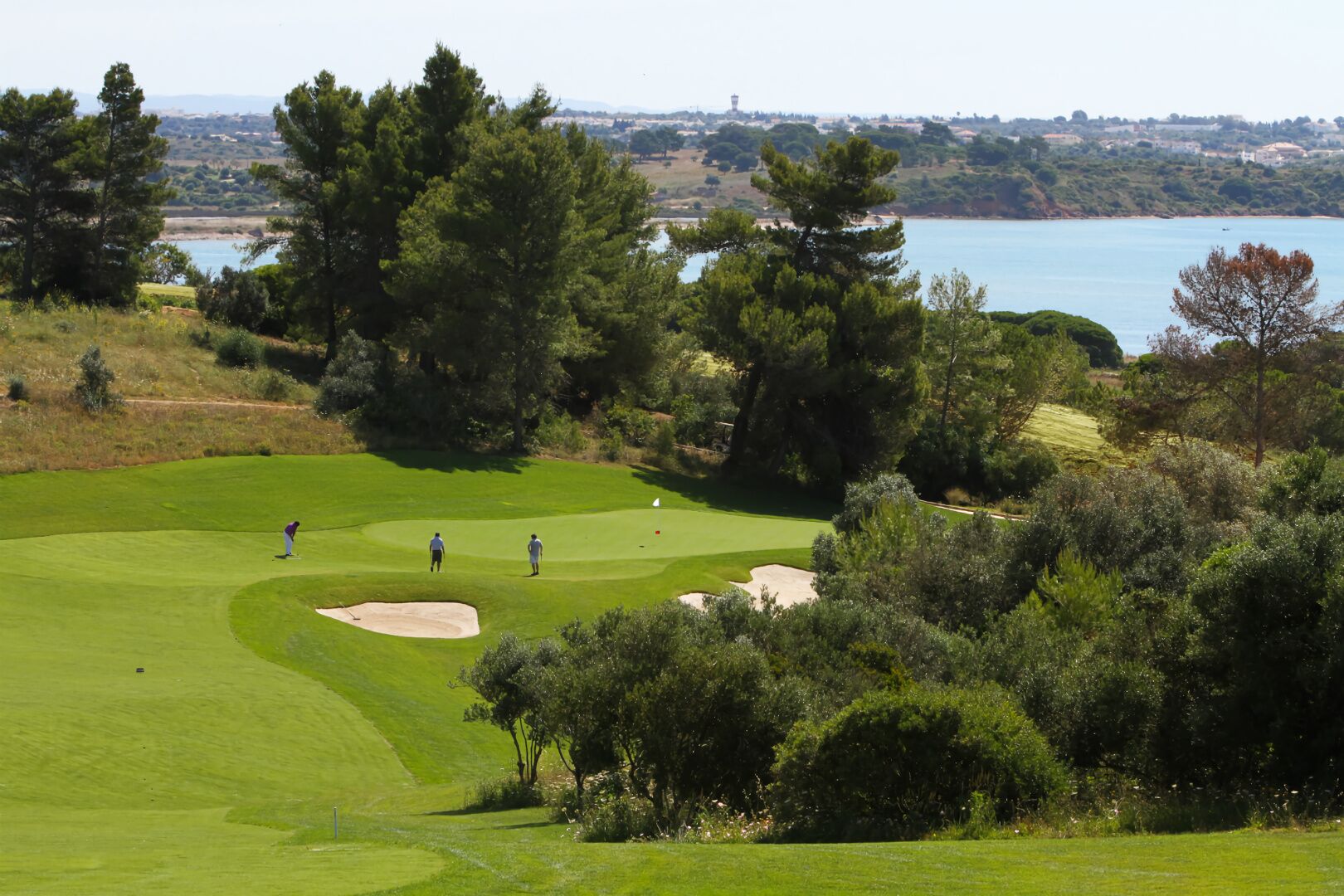 A golf course with a water body in the background, surrounded by trees and hills.