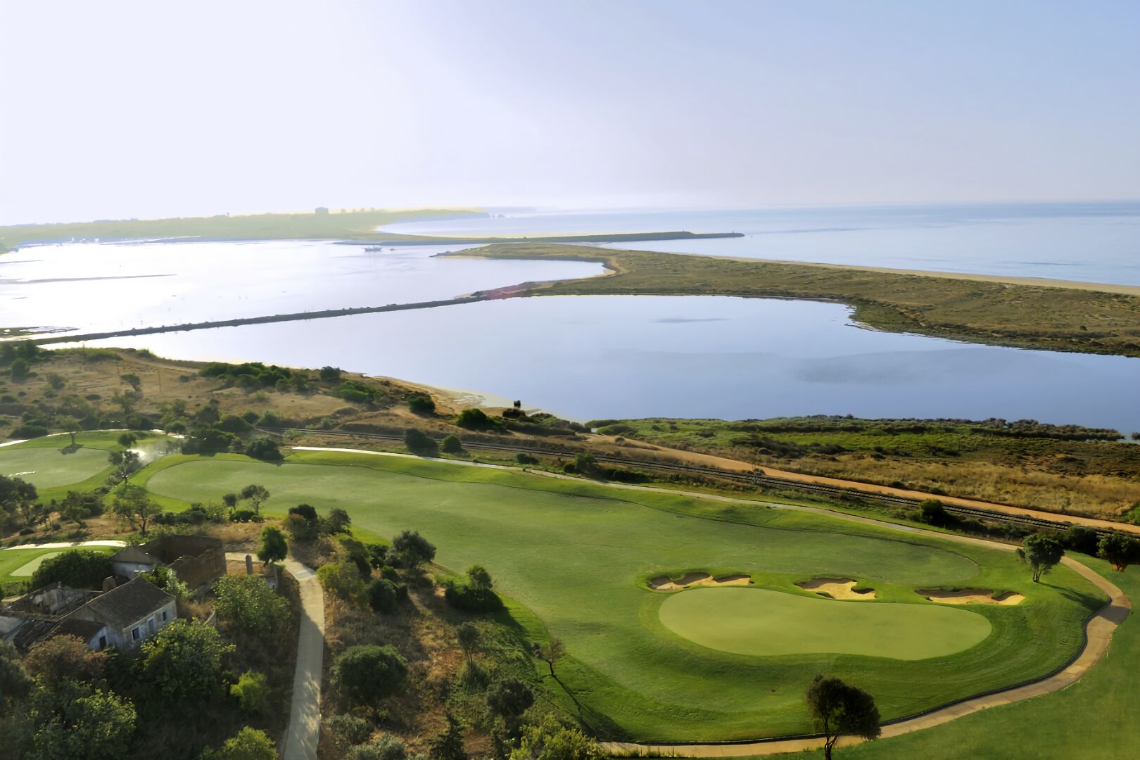 Um campo de golfe com um corpo d'água ao fundo, uma linha férrea e um edifício à esquerda.