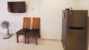 Quarto pequeno e limpo com televisão de tela plana na parede, um frigorífico Samsung, duas cadeiras de madeira e um ventilador portátil.