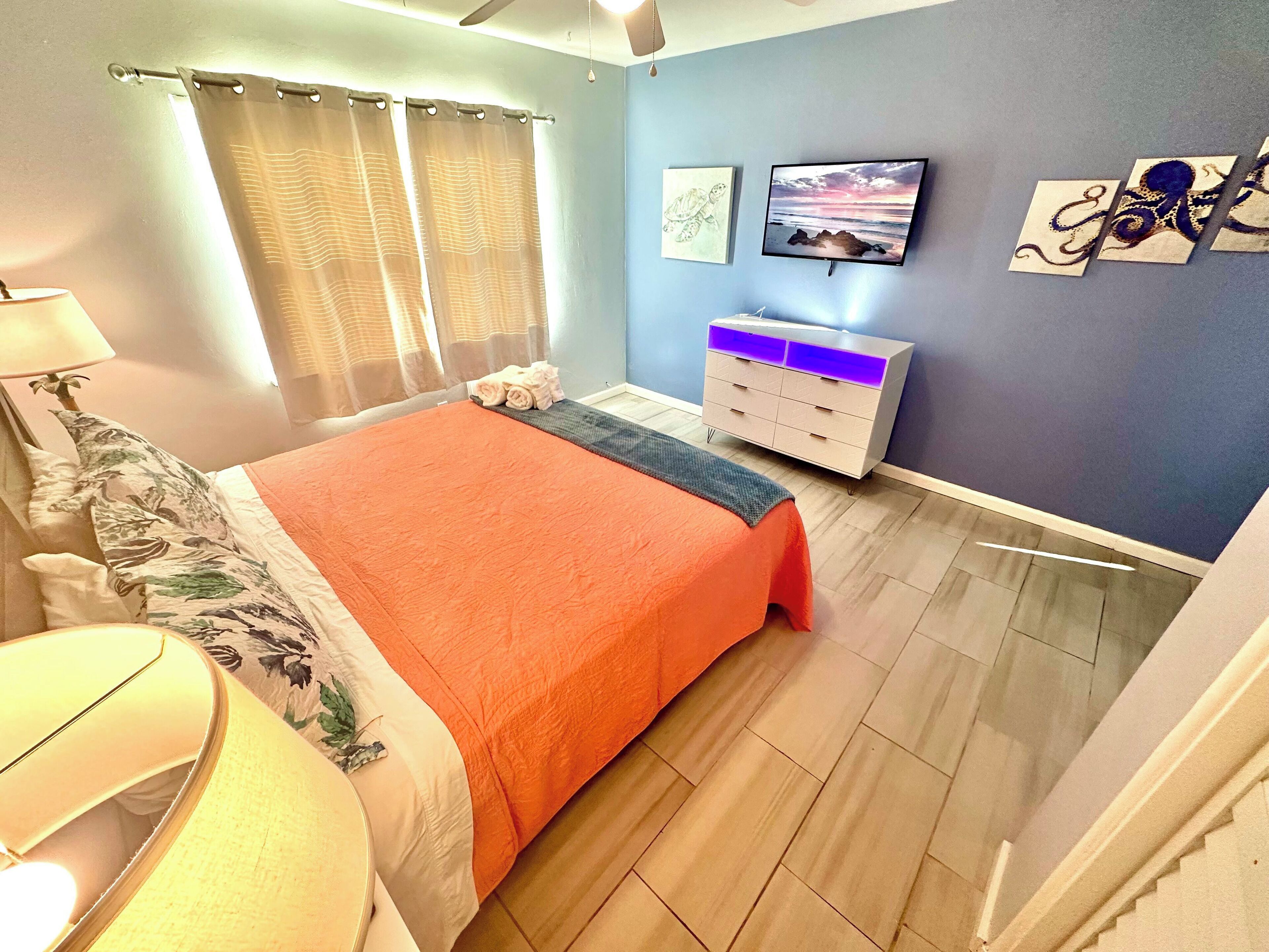 Un dormitorio con cama, mesita de noche, lámpara, cómoda y una obra de arte en la pared.