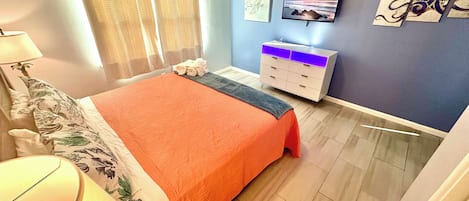 Un dormitorio con cama, mesita de noche, lámpara, cómoda y una obra de arte en la pared.