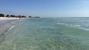 Una playa con agua clara y poco profunda, orilla de arena y algunas personas caminando.