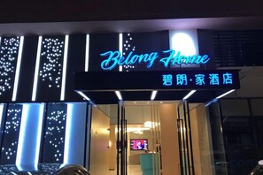 L’entrée d’un hôtel, avec une enseigne lumineuse au néon indiquant « Belong Home », et un comptoir de réception visible à l’intérieur.