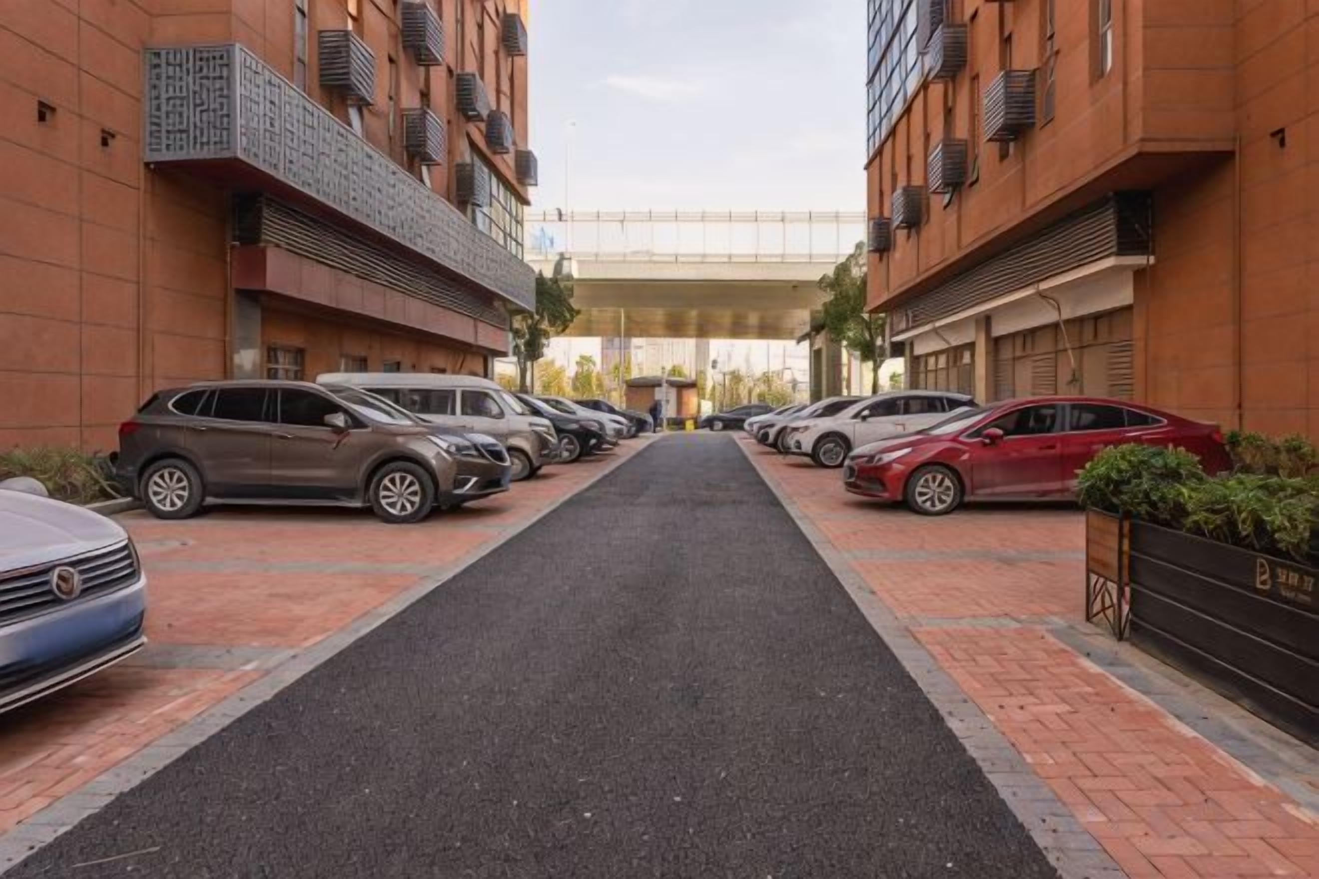 A parking area with cars parked on either side of a central path, leading to a building with a covered walkway.