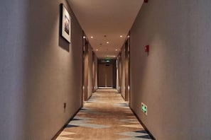 Een hotelgang met vloerbedekking, foto's in lijsten aan de muur en een brandmelder aan de rechterkant.