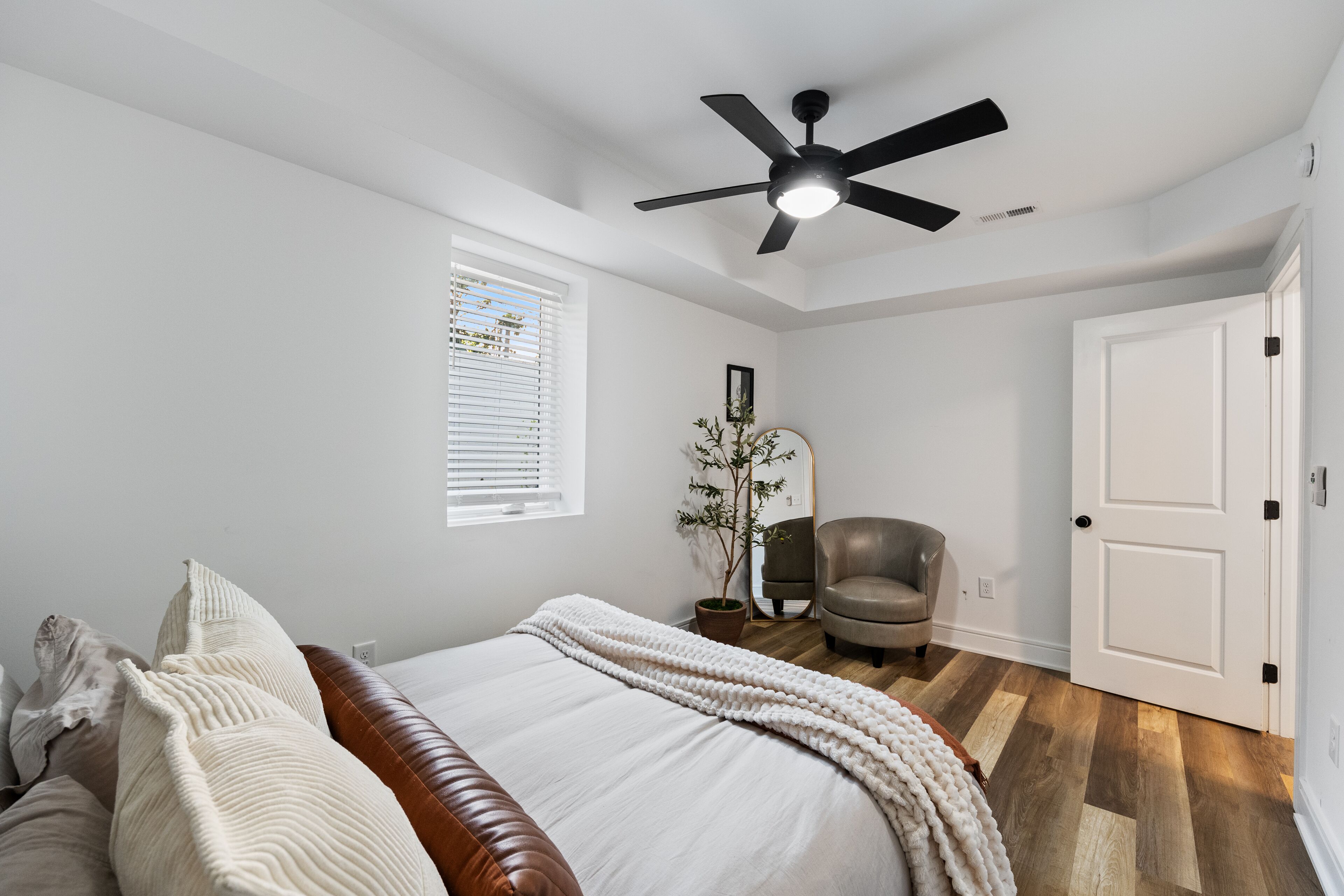 Une chambre à coucher avec un lit, un ventilateur de plafond, une fenêtre avec des stores, une plante en pot, une chaise et une porte.