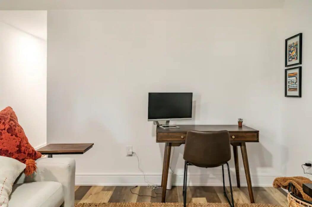 Een kamer met een houten bureau, een stoel, een televisie, een klein tafeltje en een witte bank.