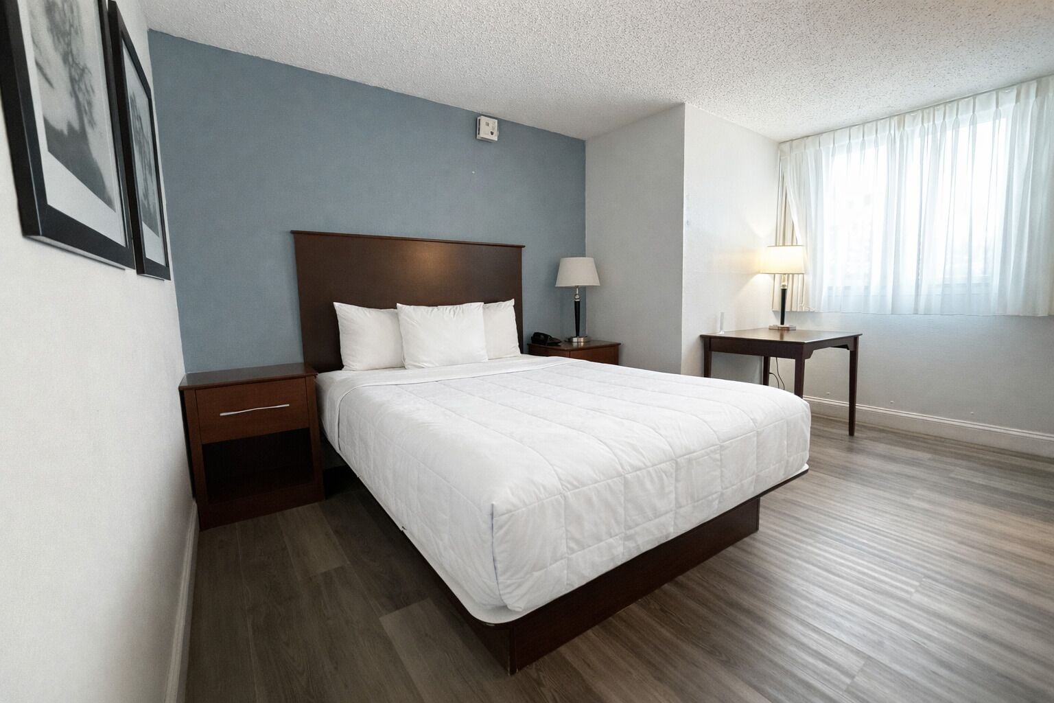 A hotel room with a large bed, wooden headboard, two bedside tables with lamps, a small desk, and a window with curtains.