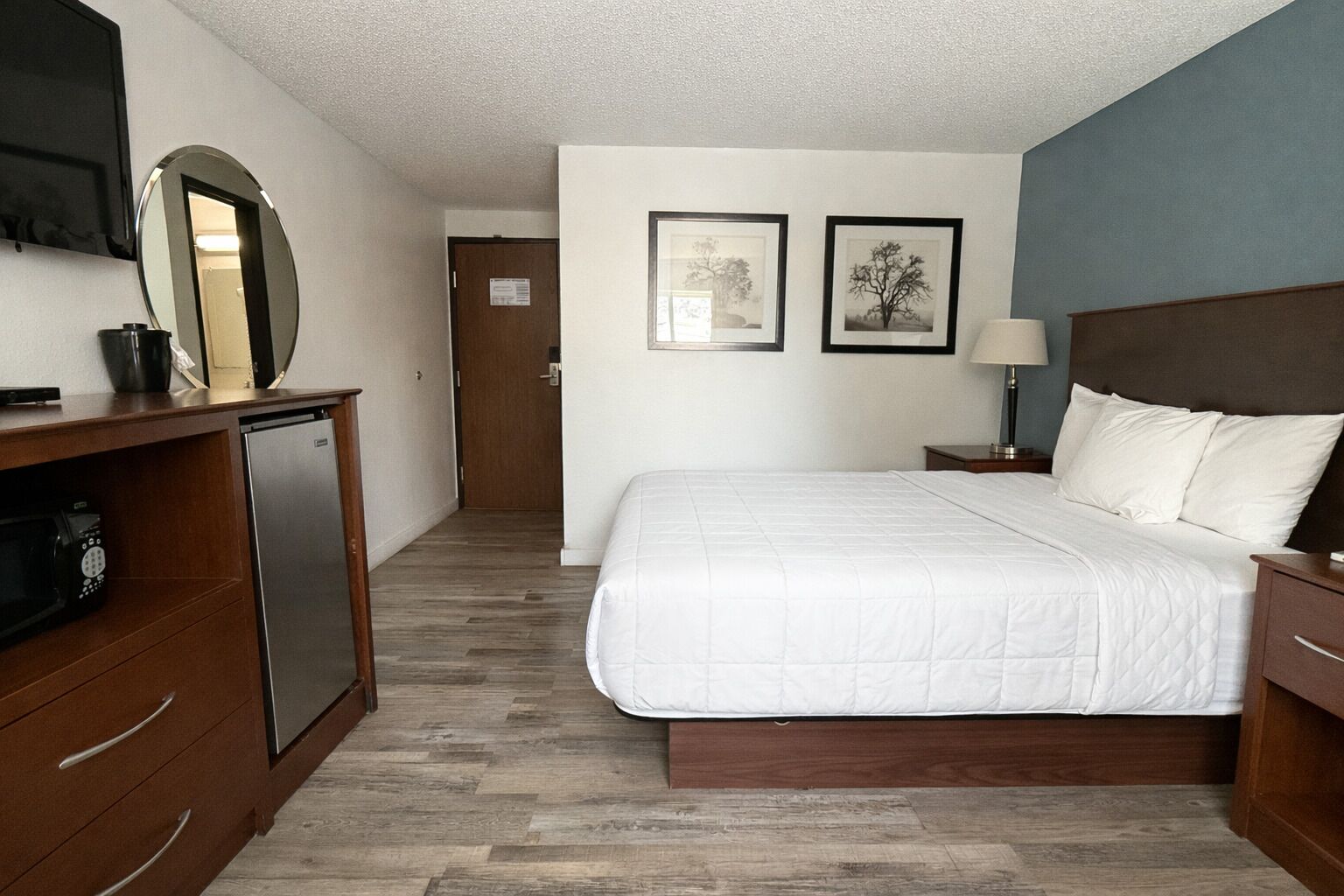 A hotel room with a bed, a desk with a computer, a mirror, and two framed pictures on the wall.