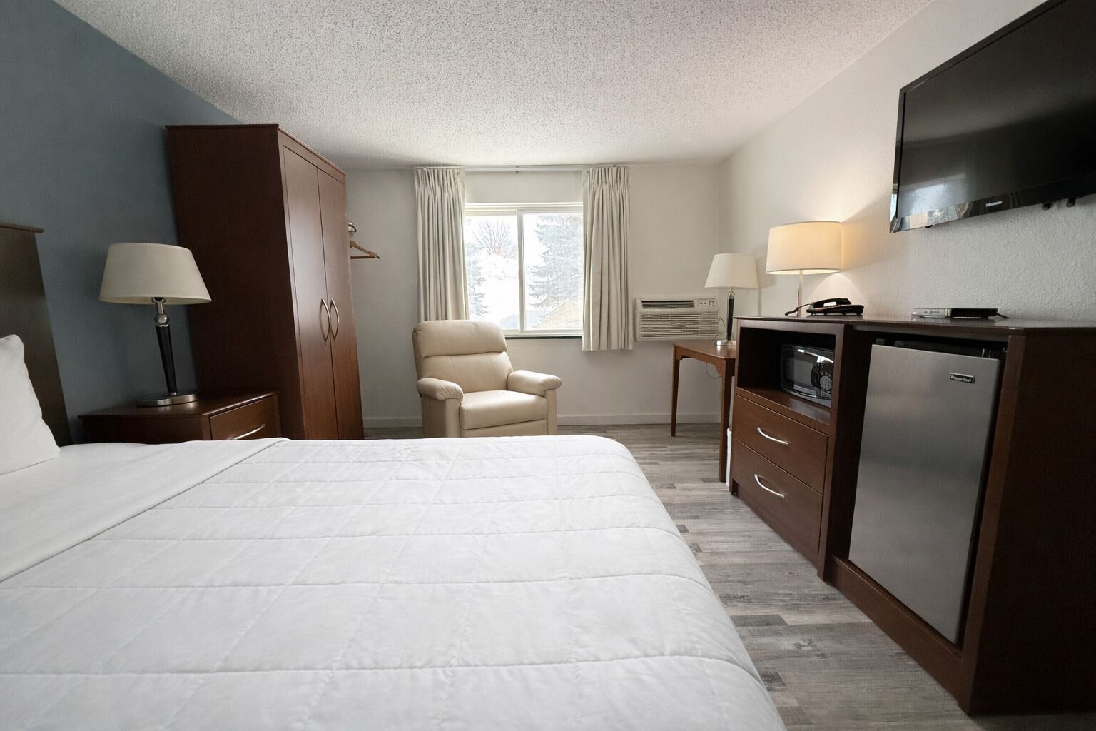 A hotel room with a large bed, a chair, a desk with a lamp, a TV, and a minibar.