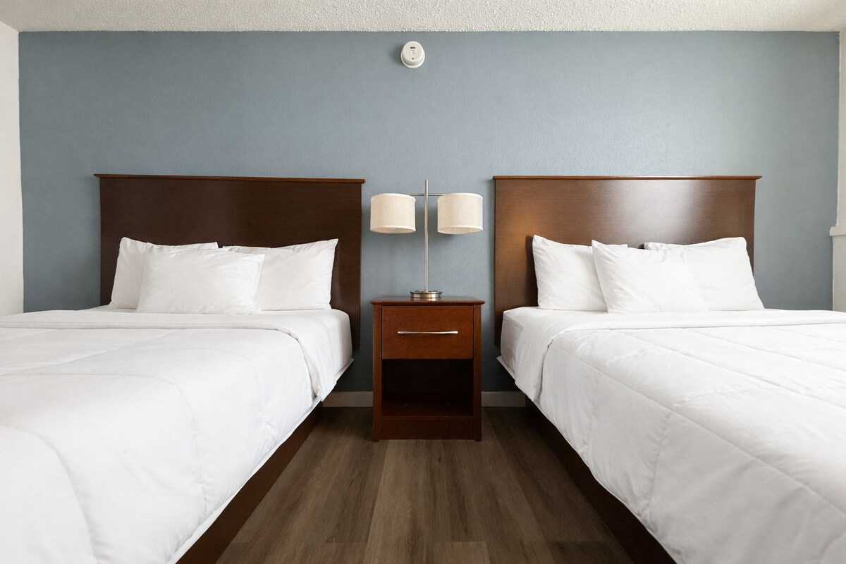 A hotel room with two beds, a nightstand with lamps, and a wall-mounted light fixture.