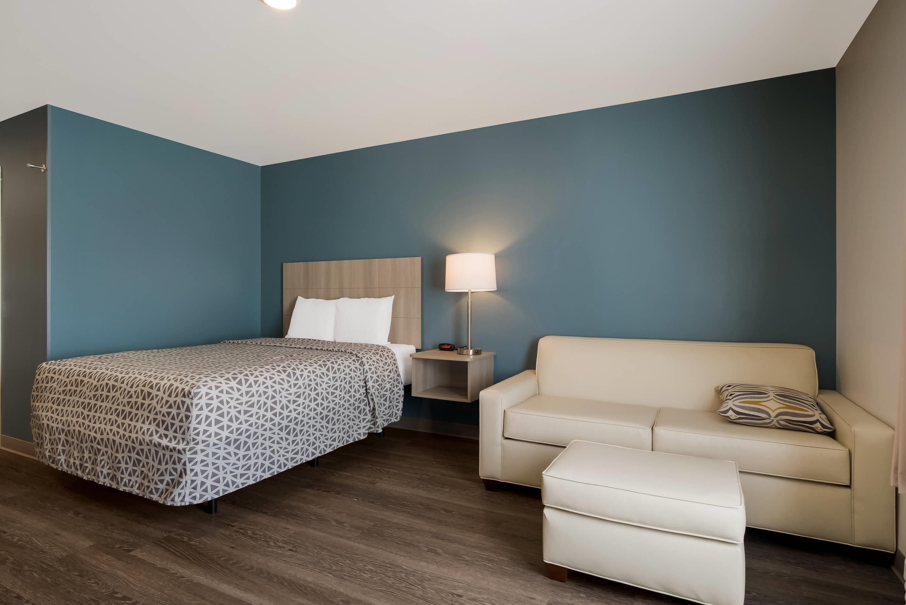 A hotel room with a bed, a nightstand with a lamp, a sofa, and an ottoman.