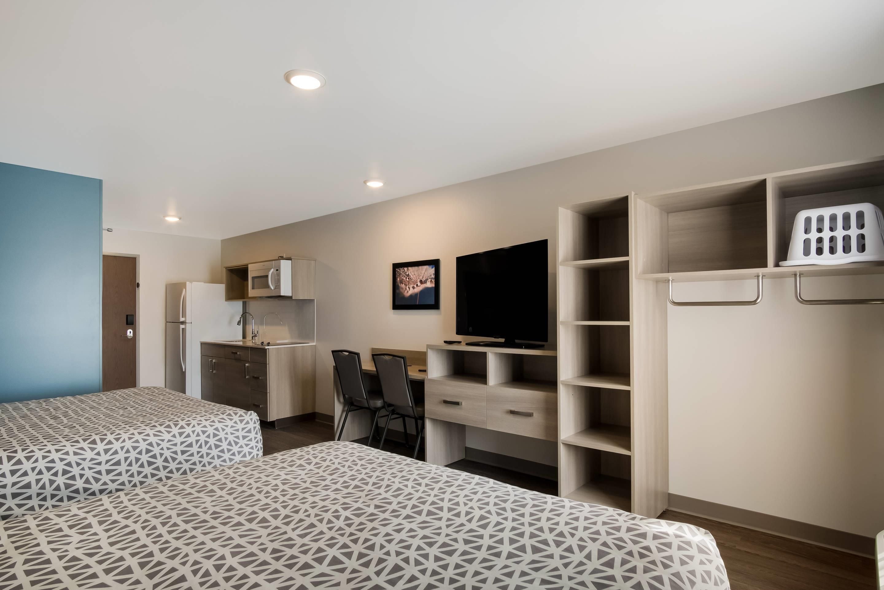 A modern hotel room with two beds, a kitchenette, a TV, and built-in storage.