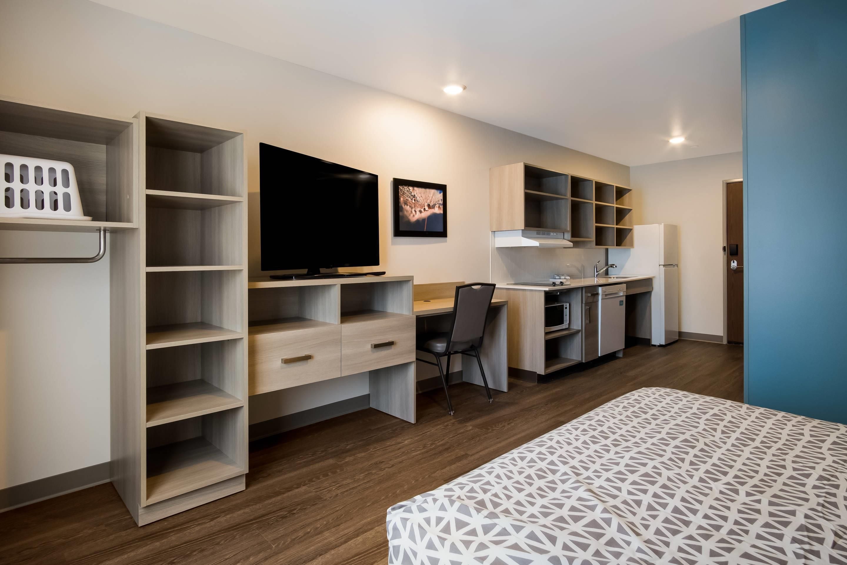 A modern hotel room with a bed, a desk, a chair, a television, and a kitchenette.
