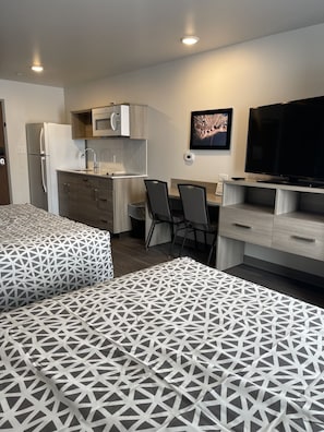A modern hotel room with a kitchenette, a flat-screen TV, two beds with geometric-patterned bedding, and a dining area. - WoodSpring Suites Grand Rapids Kentwood (Grand Rapids)