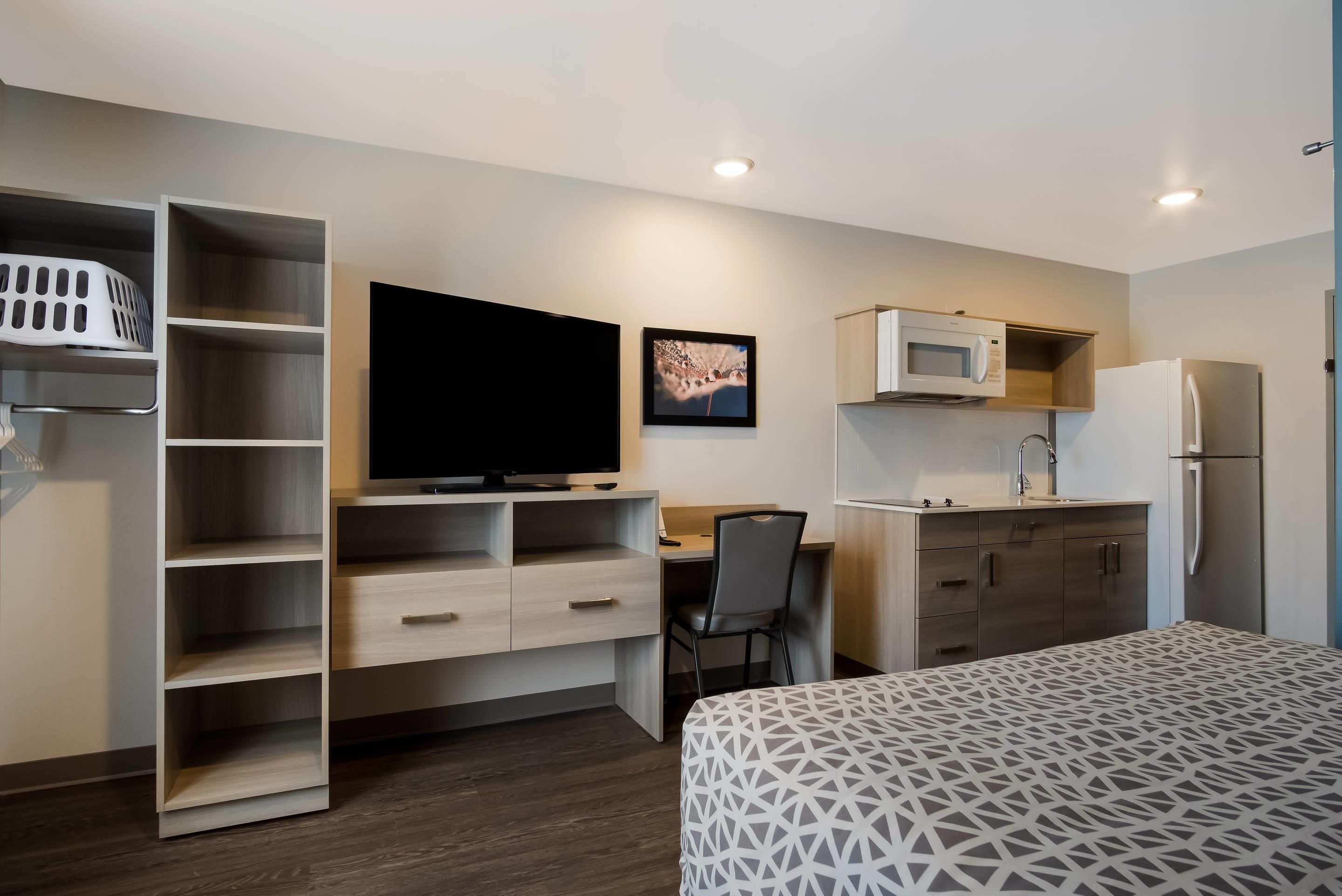 A compact hotel room with a bed, a desk with a chair, a television, a microwave, a refrigerator, and a built-in wardrobe with shelves.