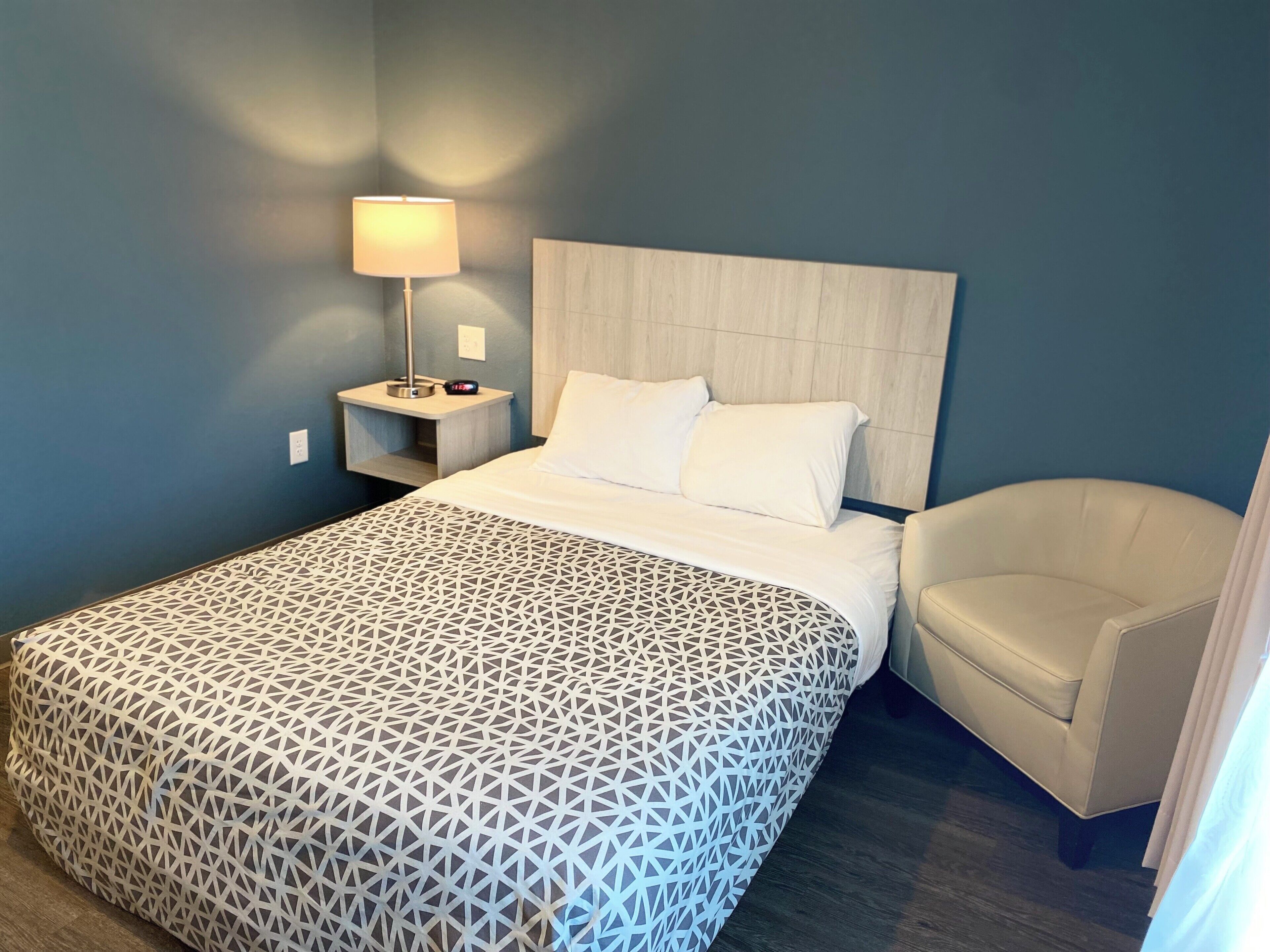 A hotel room with a bed, a nightstand with a lamp, a chair, and a patterned bedspread.