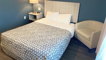 A hotel room with a bed, a nightstand with a lamp, a chair, and a patterned bedspread.