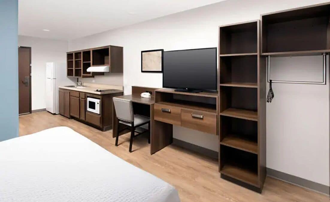 Quarto de hotel com cama, escrivaninha, televisão, armário e cozinha compacta.