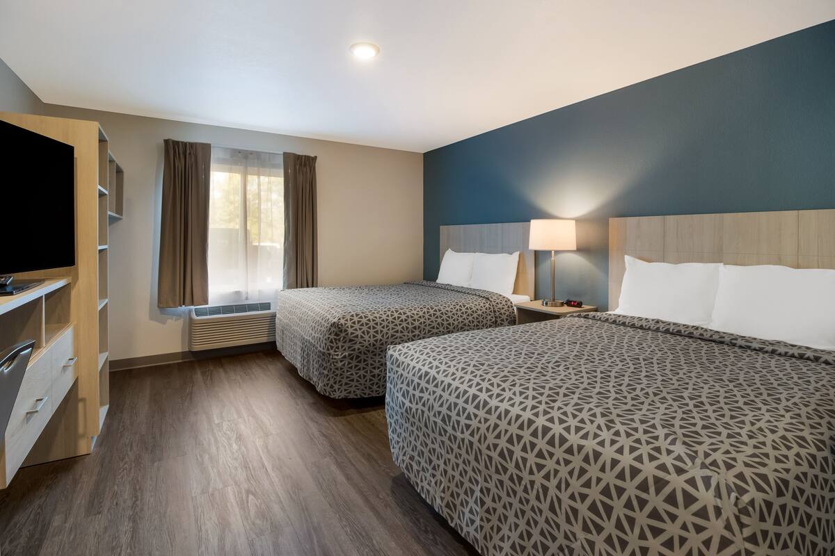Une chambre d’hôtel avec deux lits, une télévision, un bureau avec des tiroirs, une fenêtre avec des rideaux et une lampe sur la table de chevet.
