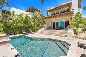 Une piscine avec des marches menant à un patio couvert, entouré de palmiers et d’un bâtiment blanc doté d’un toit en tuiles.