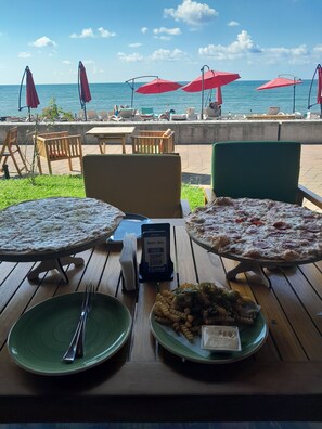 Un área de comedor al aire libre con pizzas y platos de pasta sobre mesas y sillas de madera, y sombrillas rojas.