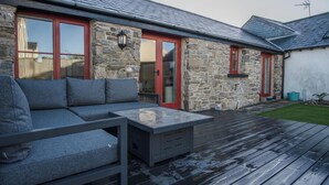 A stone house with a wooden deck, a grey sofa, and a red door. - The Old Stables - 3 Bedroom Cottage - Angle (Angle)
