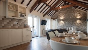 A modern kitchen with white cabinetry, a stone wall, and a dining area with a wooden table set for four. - The Old Stables - 3 Bedroom Cottage - Angle (Angle)