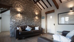 A cozy living room with a stone wall, a dark leather sofa, and a wooden ceiling. - The Old Stables - 3 Bedroom Cottage - Angle (Angle)