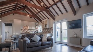 A spacious living room with a dark leather sofa, a dining area with a table and chairs, a kitchen with white cabinets and a tiled backsplash, and a large window with a view of the outdoors. - The Old Stables - 3 Bedroom Cottage - Angle (Angle)