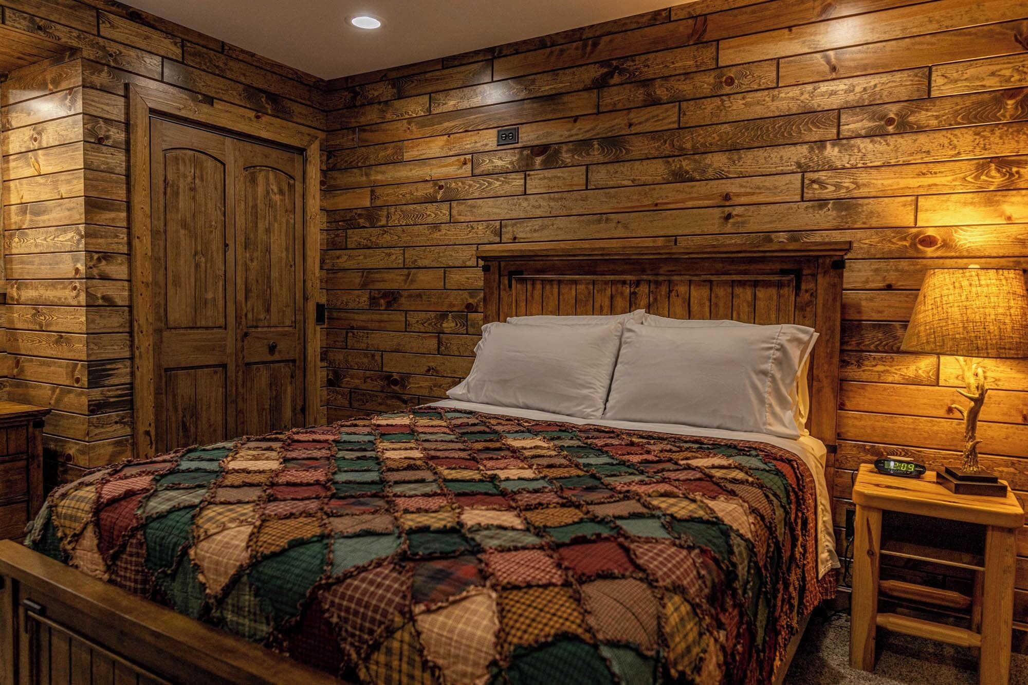 Un dormitorio con paneles de madera en la pared, una cama con colcha colorida, almohadas blancas y una mesita de noche con un reloj.
