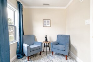 Une pièce comprenant deux fauteuils, une petite table, un tableau encadré au mur et une fenêtre avec des rideaux.