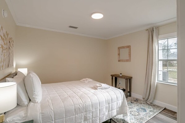 A bedroom with a bed, bedside table, lamp, and a window with curtains. - Modern Home, Great Deck & Yard- Peaceful at Potter (Mobile)
