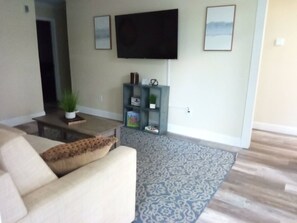 A living room with a sofa, a coffee table, a flat-screen TV mounted on the wall, a bookshelf, and a patterned rug. - Modern Home, Great Deck & Yard- Peaceful at Potter (Mobile)