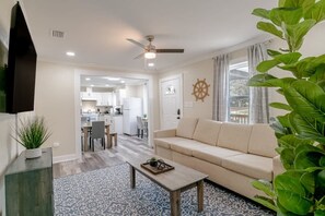 A living room with a sofa, coffee table, and a large plant. There is a ceiling fan and a view of a kitchen and dining area. - Modern Home, Great Deck & Yard- Peaceful at Potter (Mobile)