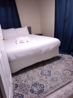 A neatly made bed with white linens and a folded towel, a small bedside table, and a patterned rug on the floor.