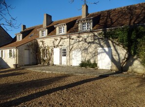 Un bâtiment traditionnel en pierre, doté de nombreuses fenêtres et portes, une cour en gravier et un arbre qui projette des ombres.