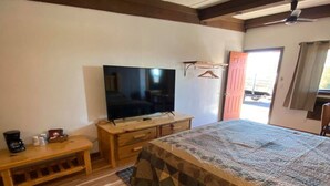 Een slaapkamer met een houten vloer, een bed met een beddengoed met een patroon, een televisie op een houten standaard, een bankje en een deur naar buiten.