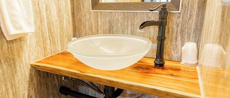 Ein Badezimmer mit einem Holzkosmetikschrank, einem Waschbecken aus Glas, einem schwarzen Wasserhahn und einem Spiegel mit strukturierter Rahmen.