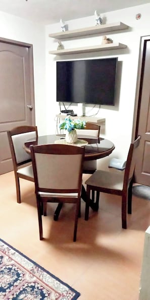 Uma área de refeições com mesa redonda, quatro cadeiras, uma TV de tela plana fixada na parede e uma pequena estante decorativa acima da TV.