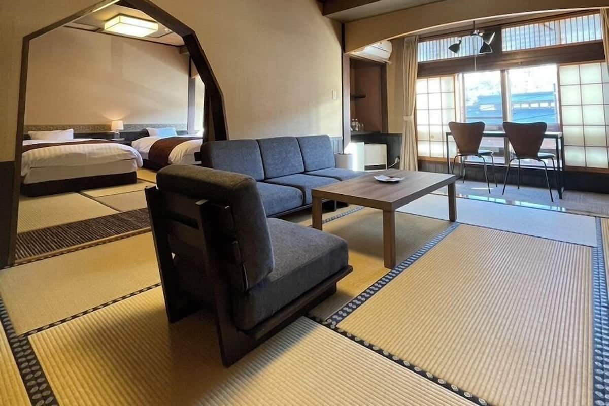A hotel room with a sofa, a small table, a bed, and a view of the outside through large windows.