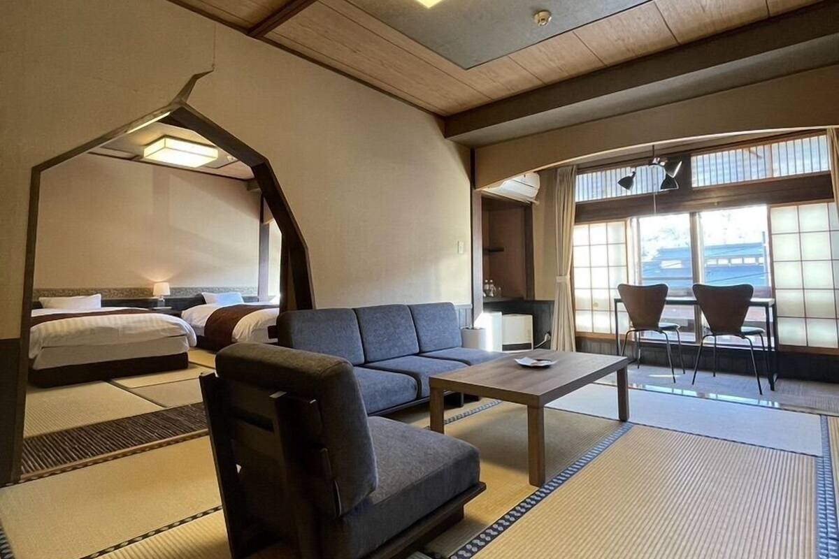 A hotel room with a sofa, a small table, a bed, and a view of the outside through large windows.